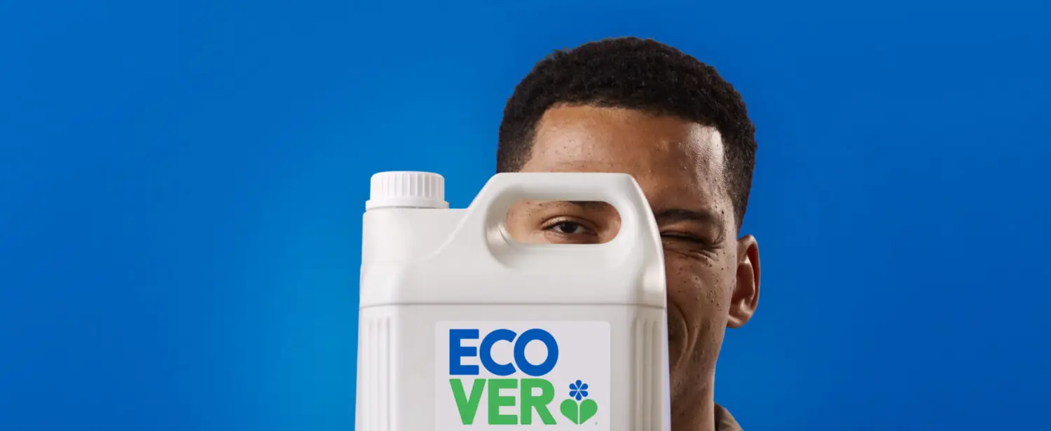 Man playfully winking while holding a large Ecover cleaning product bottle in front of his face against a blue background.