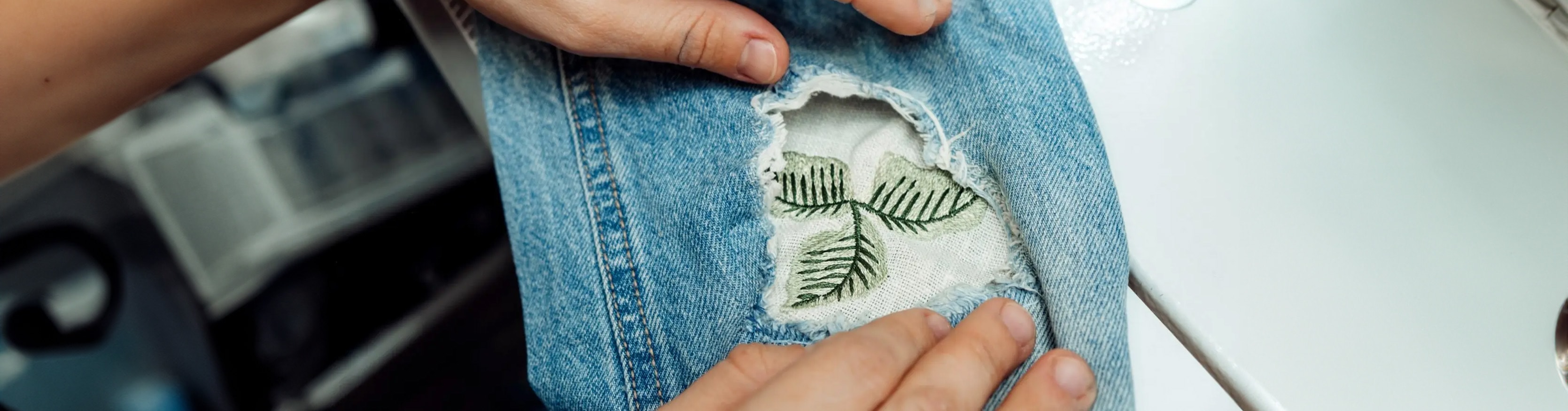 Hands holding blue jeans with a ripped section revealing fabric with embroidered green leaves, near a sewing machine.